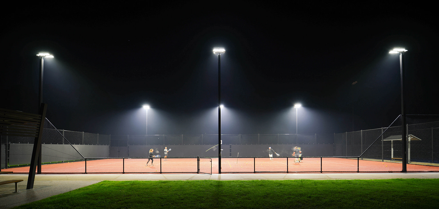 Night view of Eltham Tennis Club illuminated by multi-court LED lighting, with players on the court demonstrating the even light distribution and visibility.